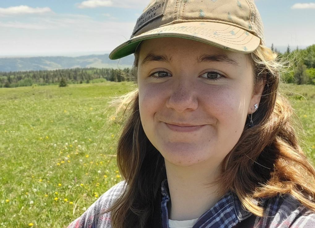 DarynFarrant_CNN_IntroBlog A photo of Daryn: A woman with shoulder-length wavy light brown hair and brown eyes, wearing a ball cap with trees on it and a blue plaid shirt. A pair of binoculars is slung across her chest.