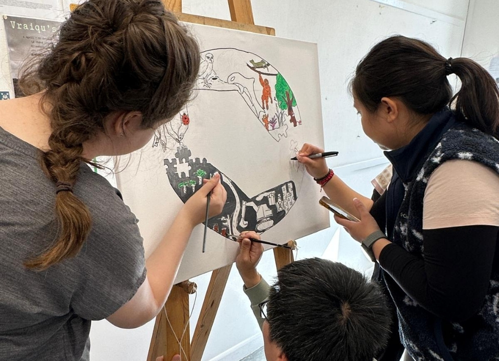 Daryn and her two classmates all draw on a white canvas leaning on an easal. The three are working together to create a circular drawing.
