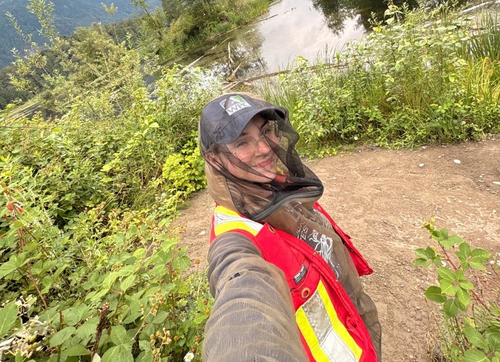 A biologist with a high visibility vest and but netting stands next to a marshy area. She smiles at the camera, taking a selfie.