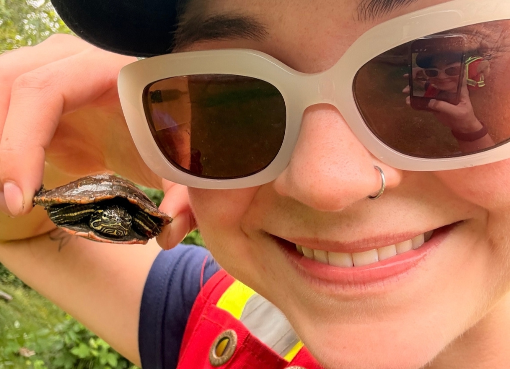 A biologist holds up a small painted turtle nest to her face, and smiles. You can see her cellphone in the reflection of her sunglasses.