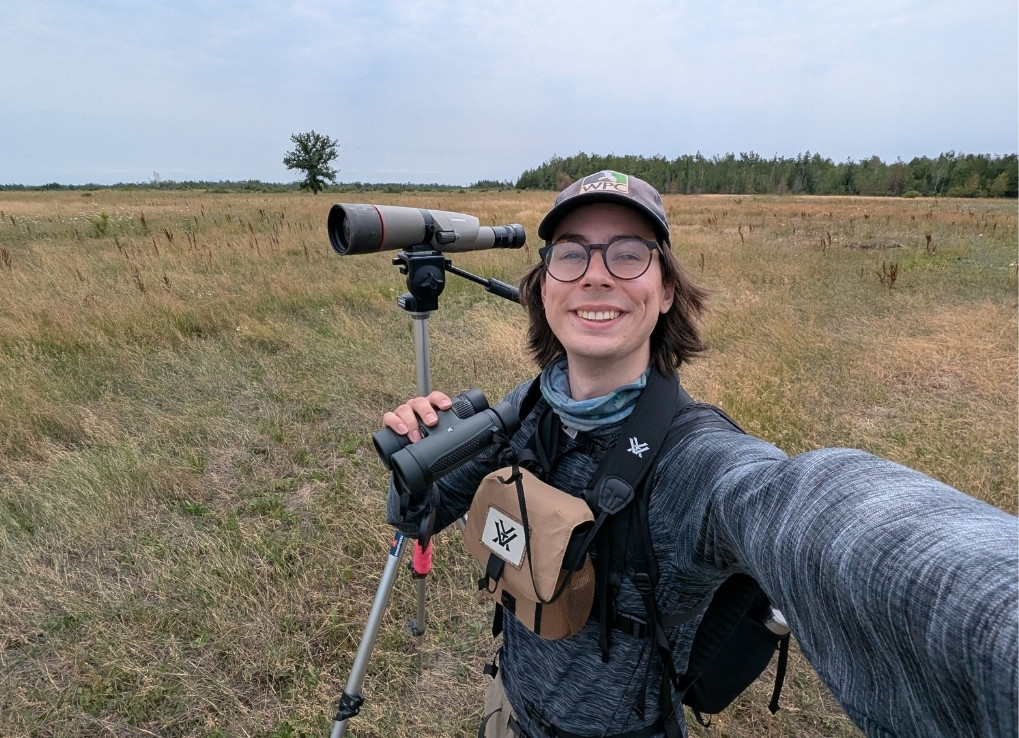 A biologist stands in a brown, grassy field with a scope and binoculars. They smile at the camera.