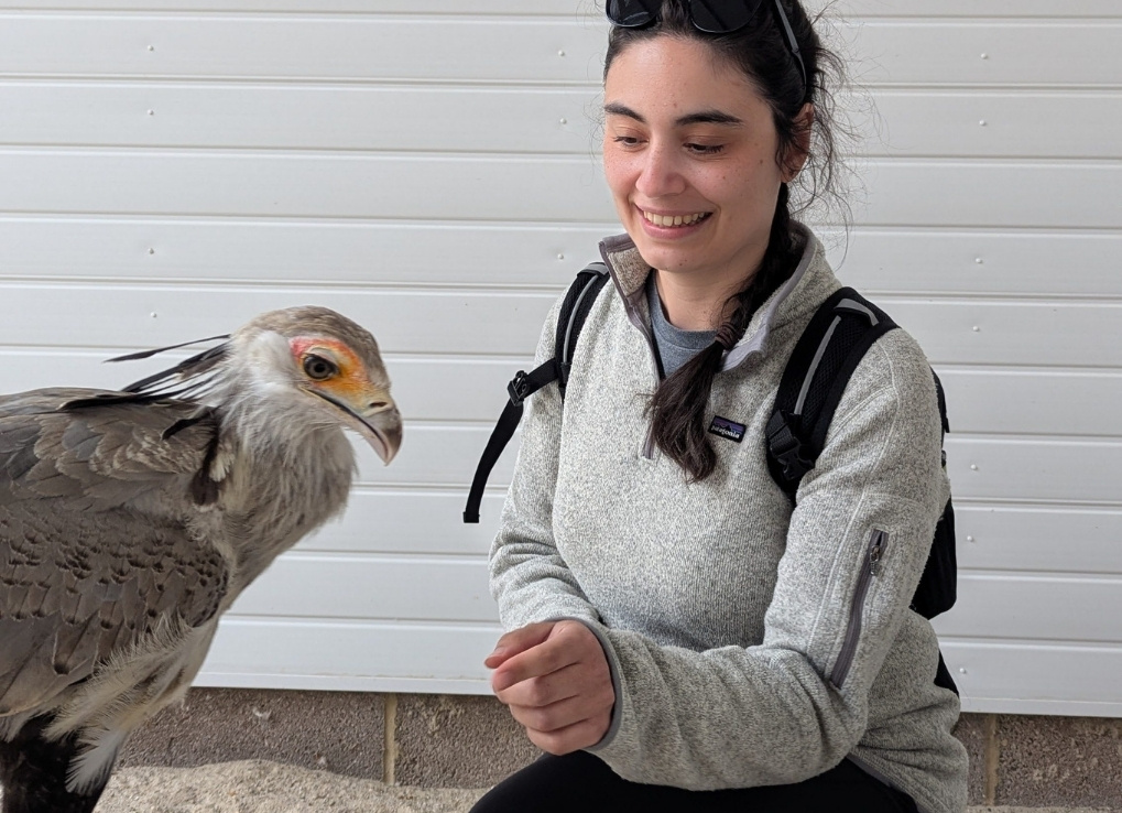 Jenna squats, reaching her hand out to a white bird with an orange ring around its eye and large black head feathers.