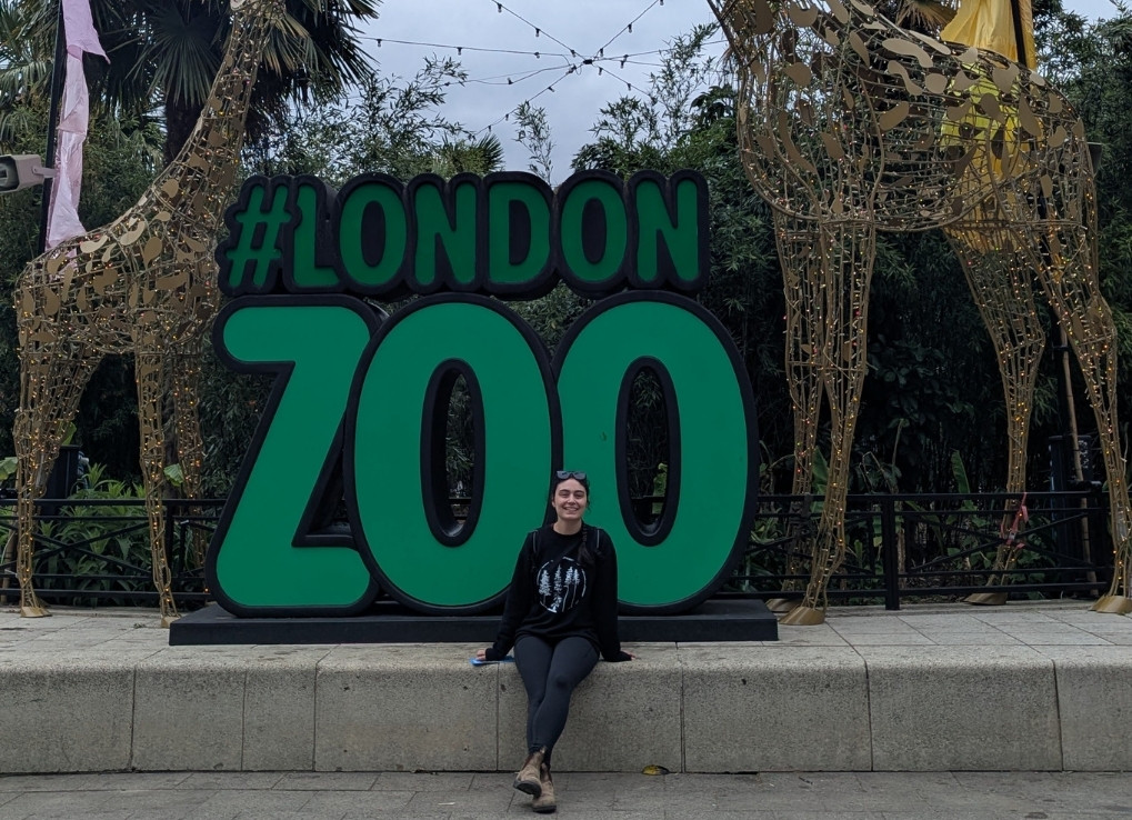 Jenna sits at the base of a very large green sign that says #LondonZoo. The sign is located between two metal giraffe wire-like statues.